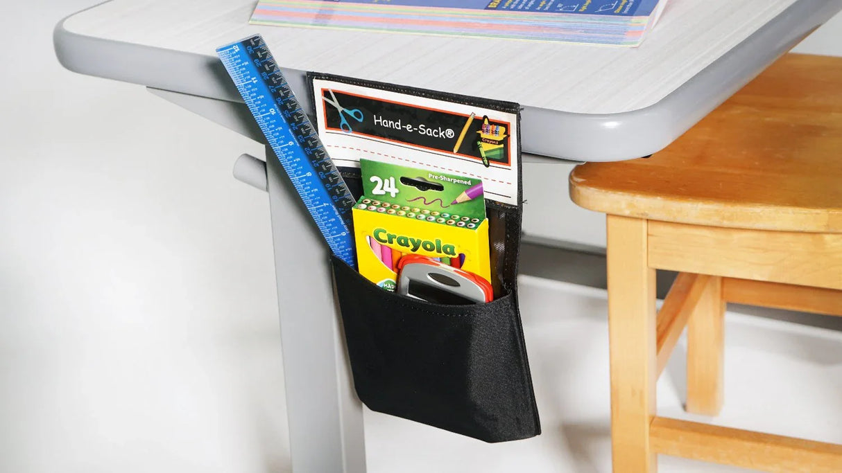 Black Hand-e-Sack attached to school desk, holding pencils, ruler, and stationery supplies.