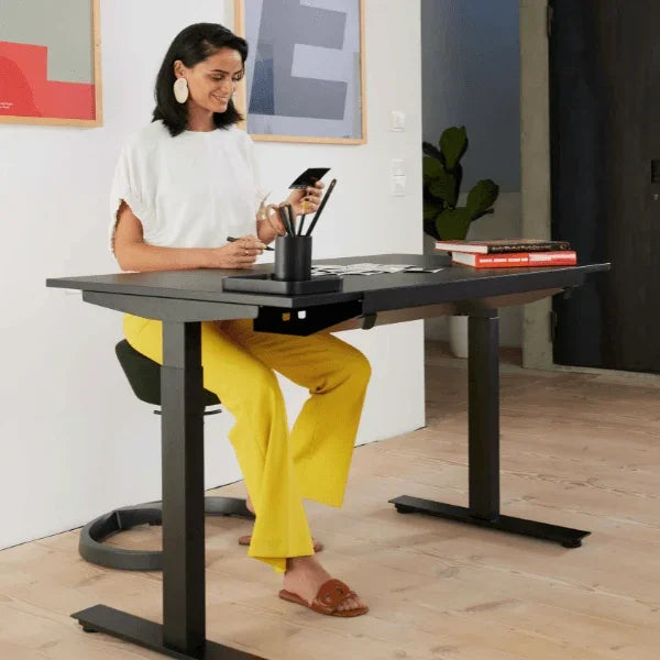 Woman sitting on an Aeris Swopper ergonomic office stool at a black standing desk.