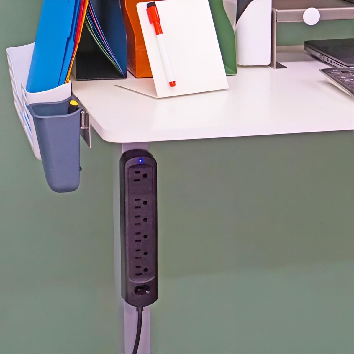 A black multi-outlet power strip mounted vertically on the side of a white desk against a sage green wall, with office supplies including colorful folders, a white notepad holder with a red pen, and a monitor nearby.
