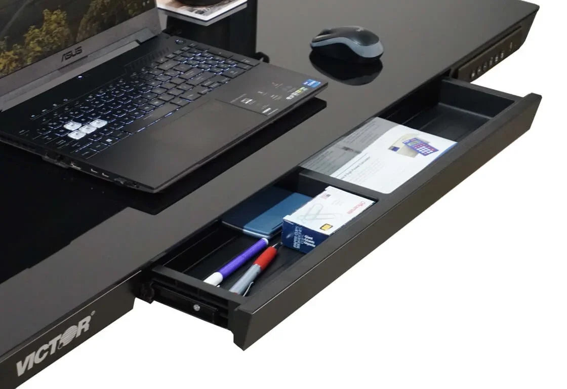 Victor® Electric Glass Topped Standing Desk with open under-desk storage drawer holding office supplies.