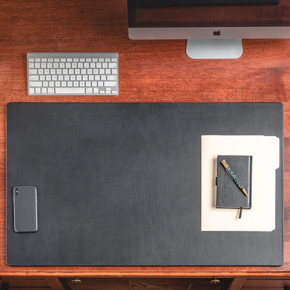 Black leather desk mat on wooden desk with phone, folder, notebook, pen, keyboard and monitor