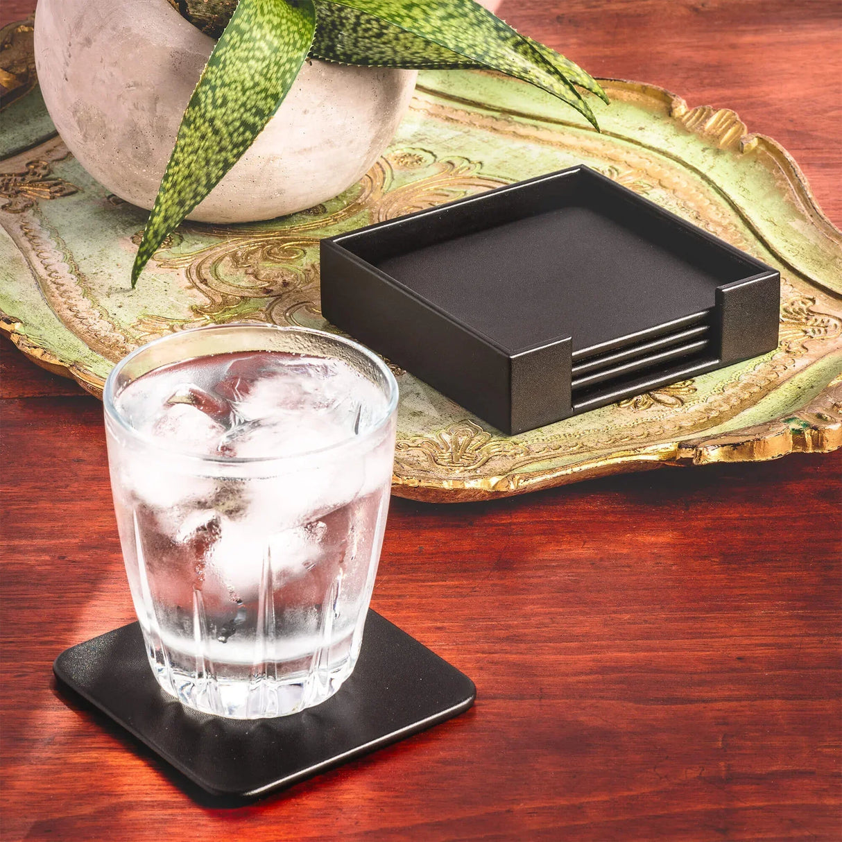 Black leather square coaster set with holder on wooden table, glass of ice water on coaster