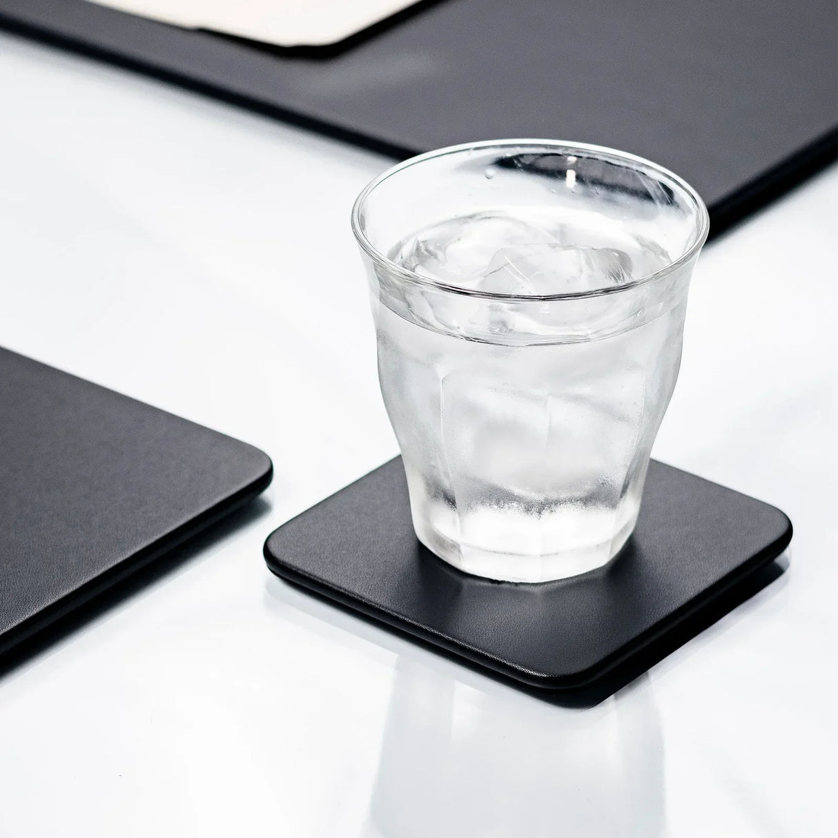 Glass of iced water on a black leatherette square coaster on a white desk