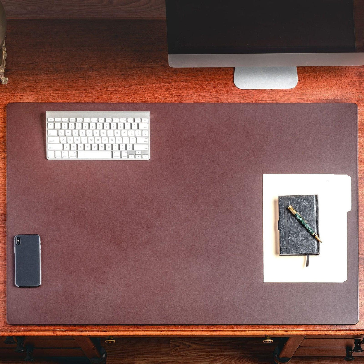 Chocolate brown leather desk mat on wooden desk with keyboard, phone, and notebook setup