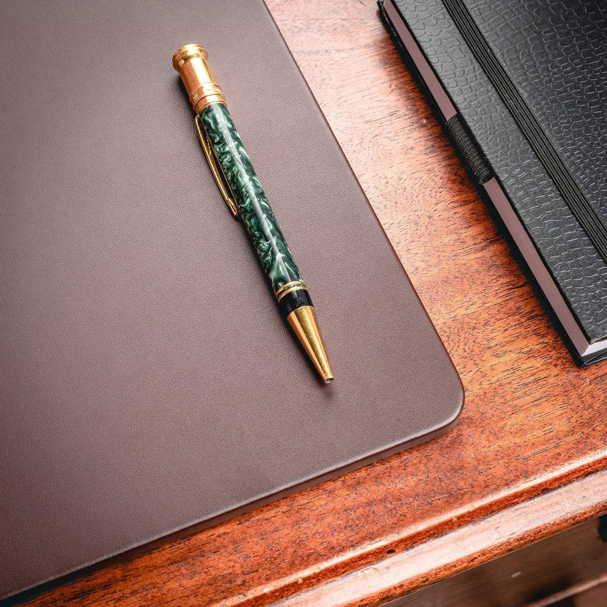 Chocolate brown leather desk mat on wooden desk with a green pen and black notebook