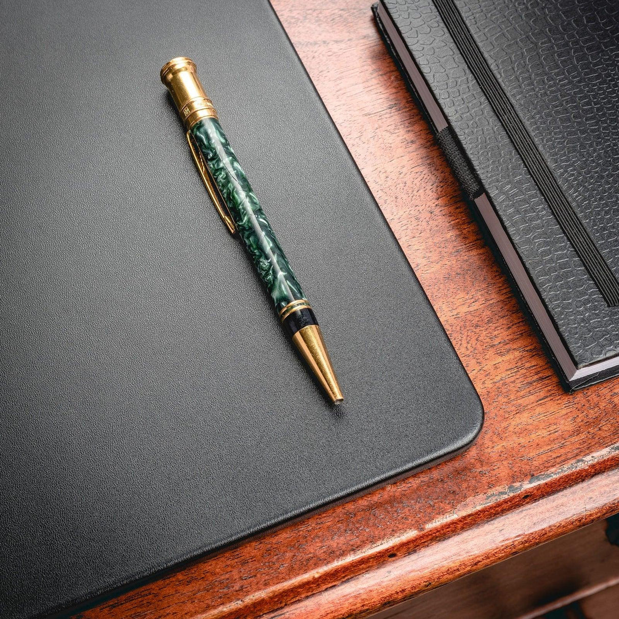 Black leather desk mat on wooden desk with green and gold pen and textured notebook