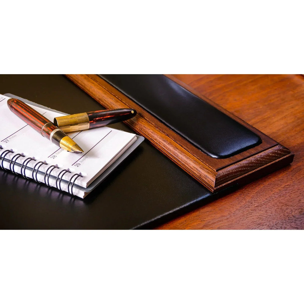 Walnut and black leather desk pad with side rail, notebook, and pens on wood desk