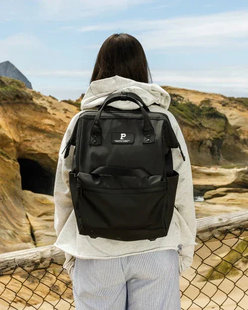 Person wearing black backpack with P logo, standing by coastal cliffs, ideal for school or office essentials