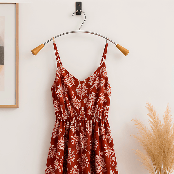 A rust red floral spaghetti-strap dress hanging on a curved metal hanger with wooden tips against a white wall, with a framed print and dried pampas grass partially visible in the background.