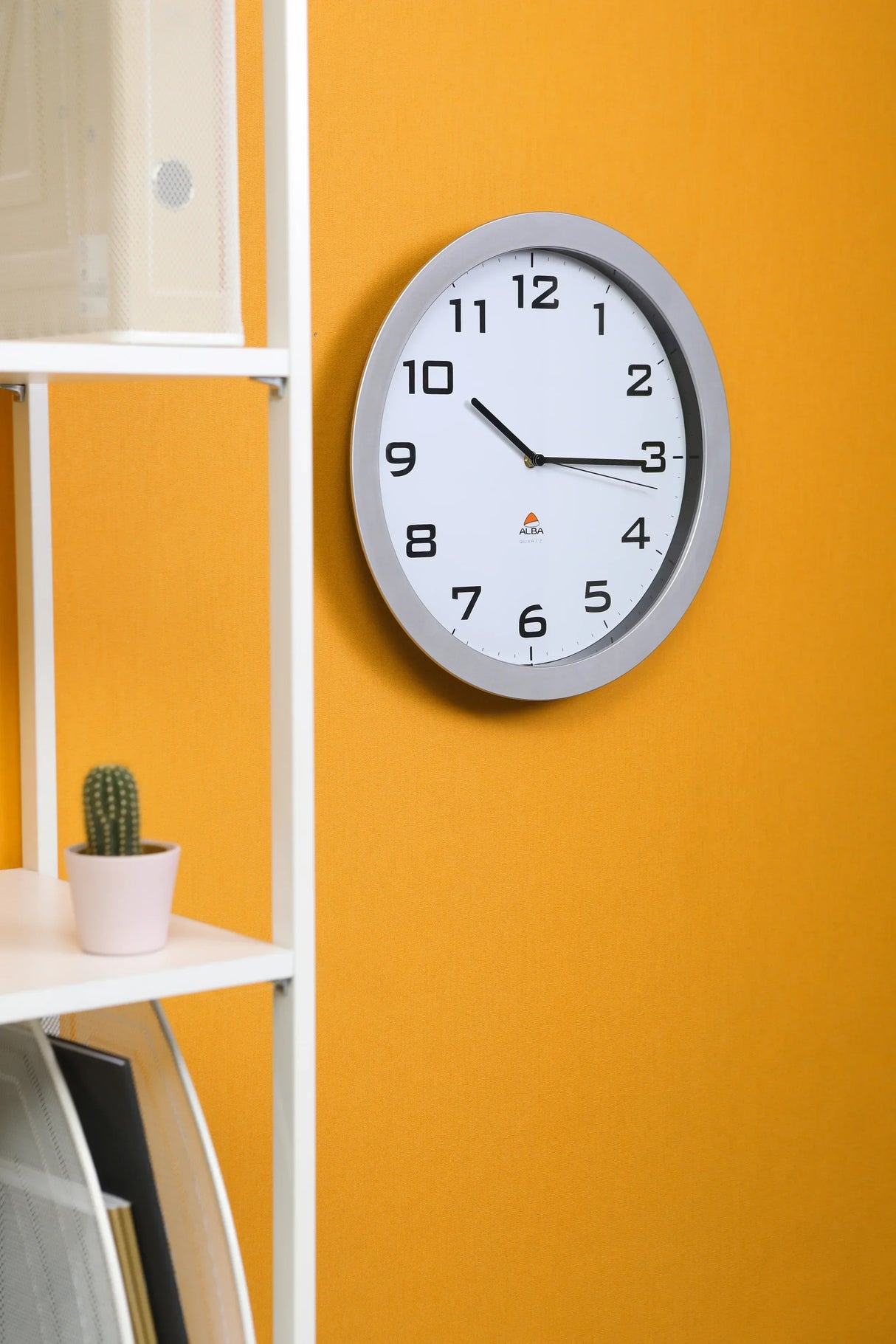 Modern wall clock with silver frame on orange wall, beside white shelving with cactus, ideal for office or school organization.