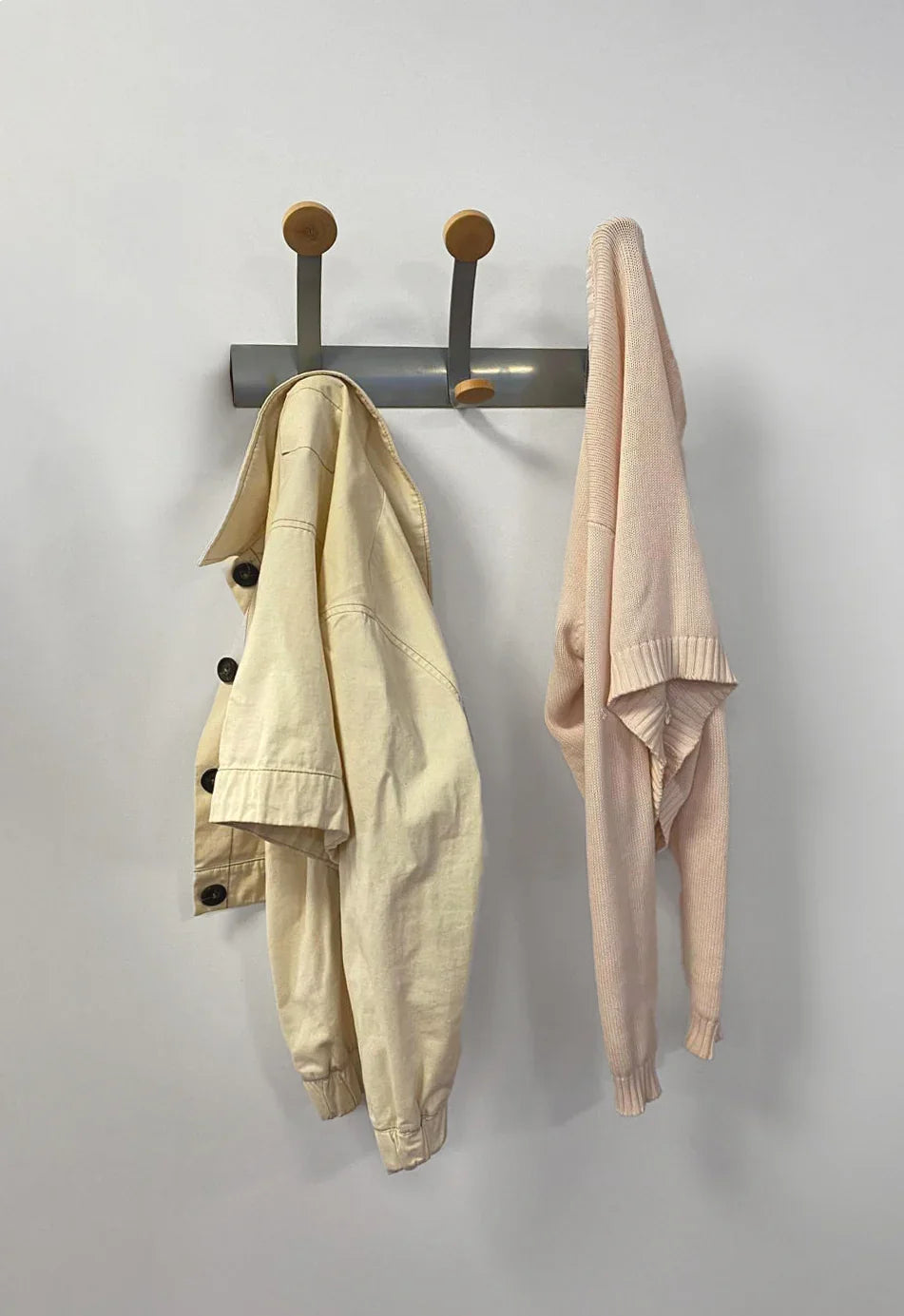 Wall-mounted coat rack with wood pegs holding a beige jacket and light pink sweater