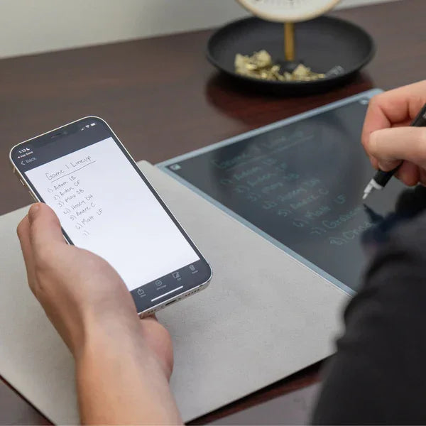 Person writing notes on a smart notebook while referencing a notes app on a smartphone at desk.