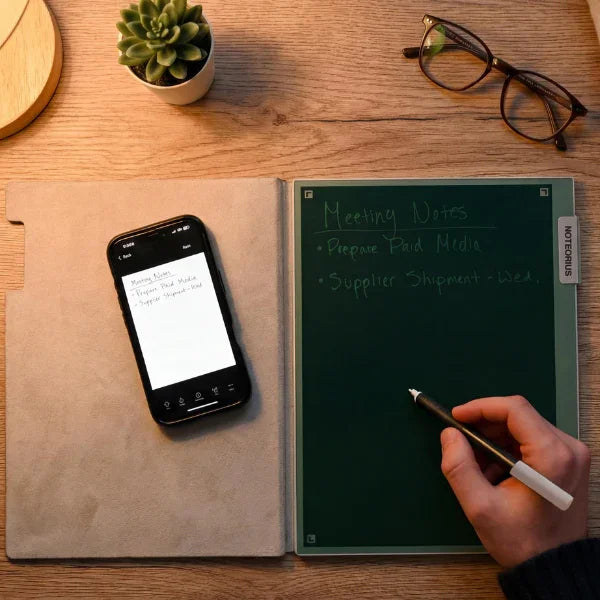 Person writing meeting notes in smart digital notebook with smartphone, glasses, and plant on desk
