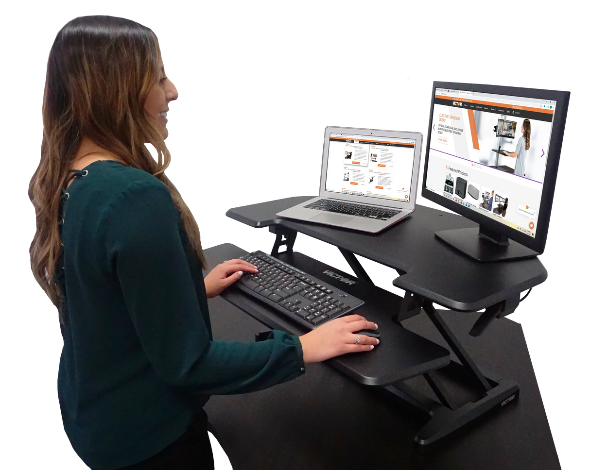 Woman using high-rise adjustable corner standing desk with monitor, laptop, and keyboard tray