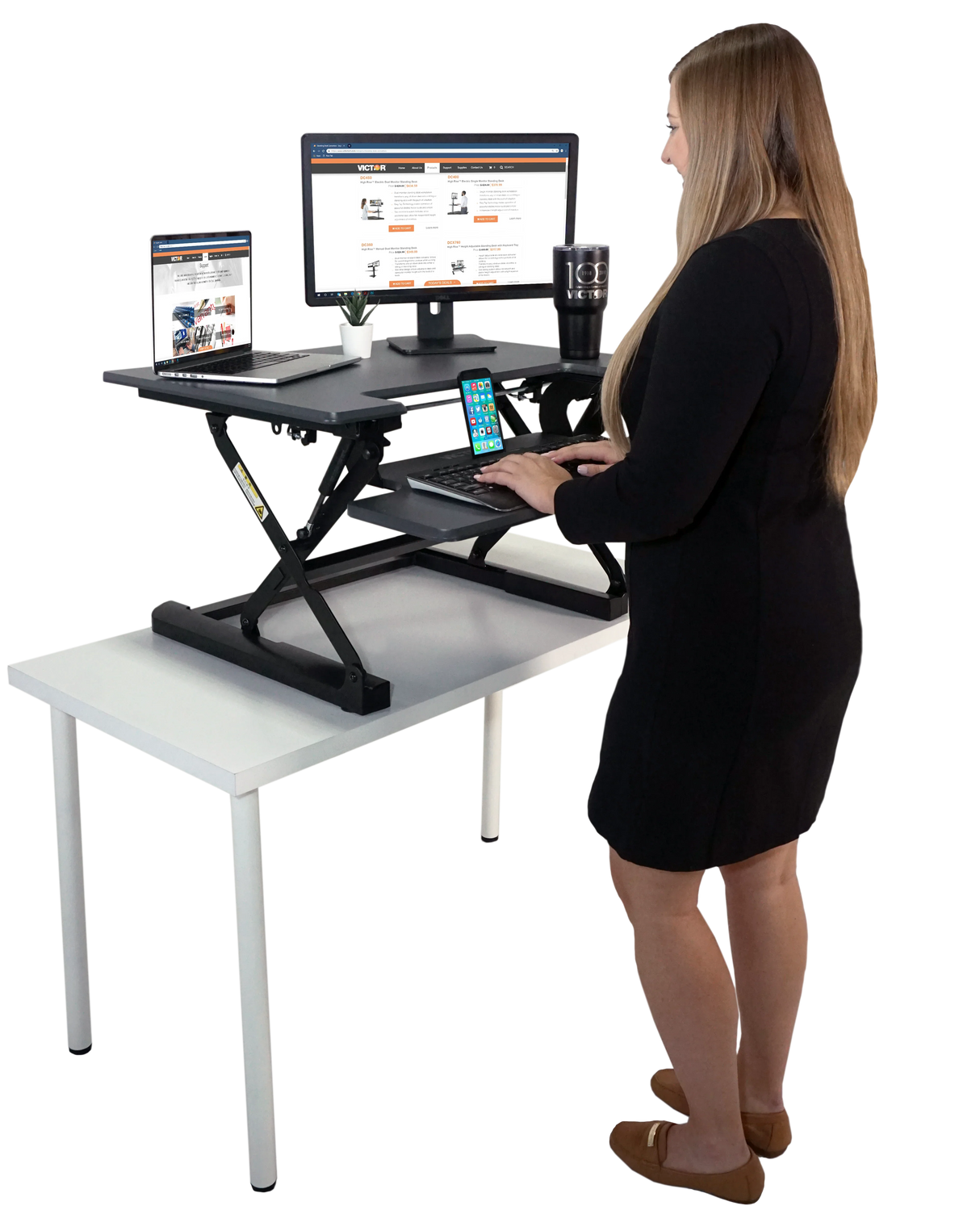Woman using black adjustable standing desk converter with monitor, laptop, and smartphone on white table