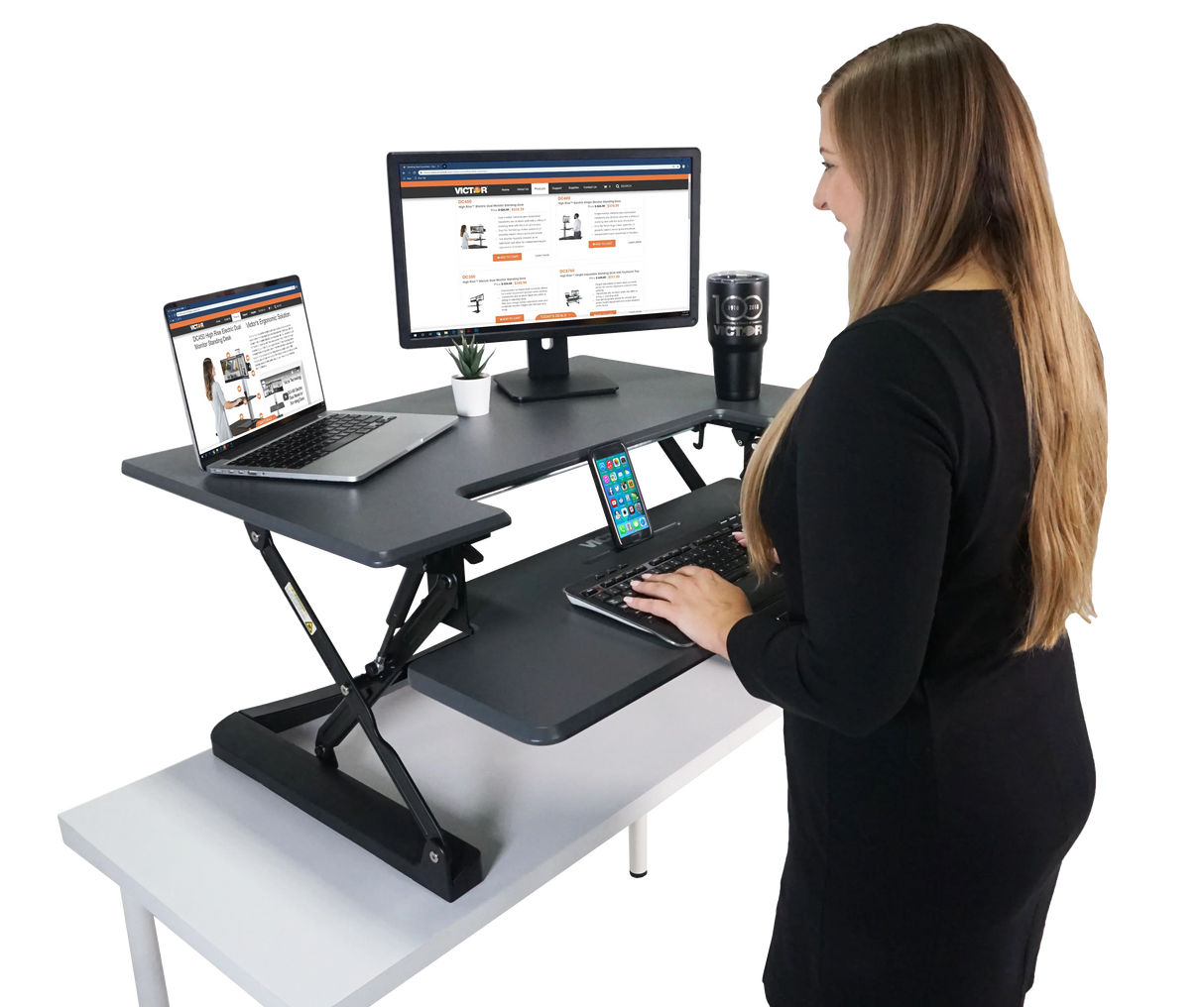 Woman at adjustable standing desk with monitor, laptop, keyboard tray, and phone holder