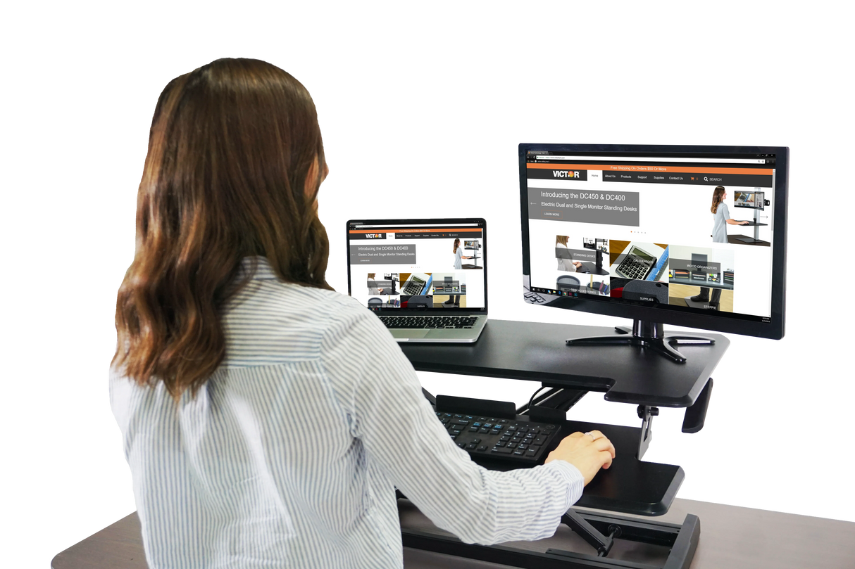 Woman using adjustable height standing desk with monitor, laptop, and keyboard tray in office