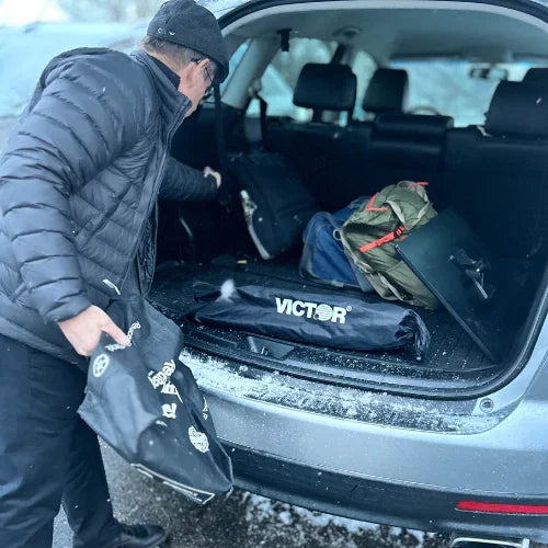 Person loading Victor-branded portable standing desk bag and gear into car trunk on snowy day