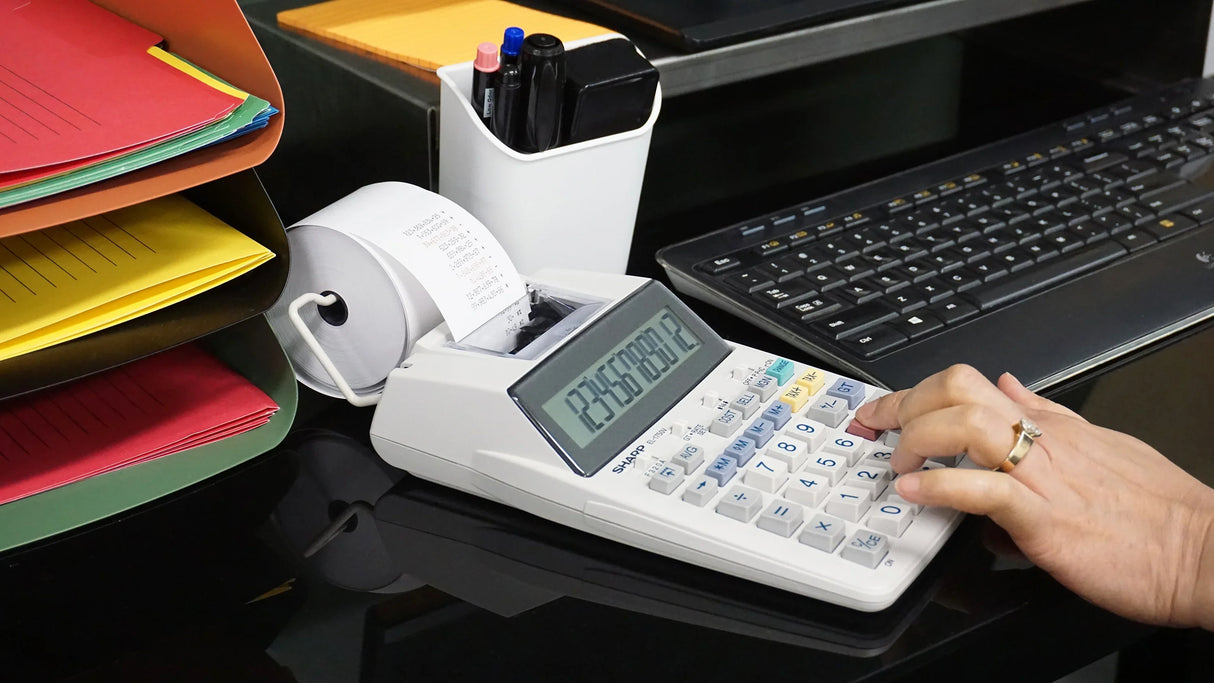 Hand using desktop printing calculator on office desk with keyboard, files, and organizer