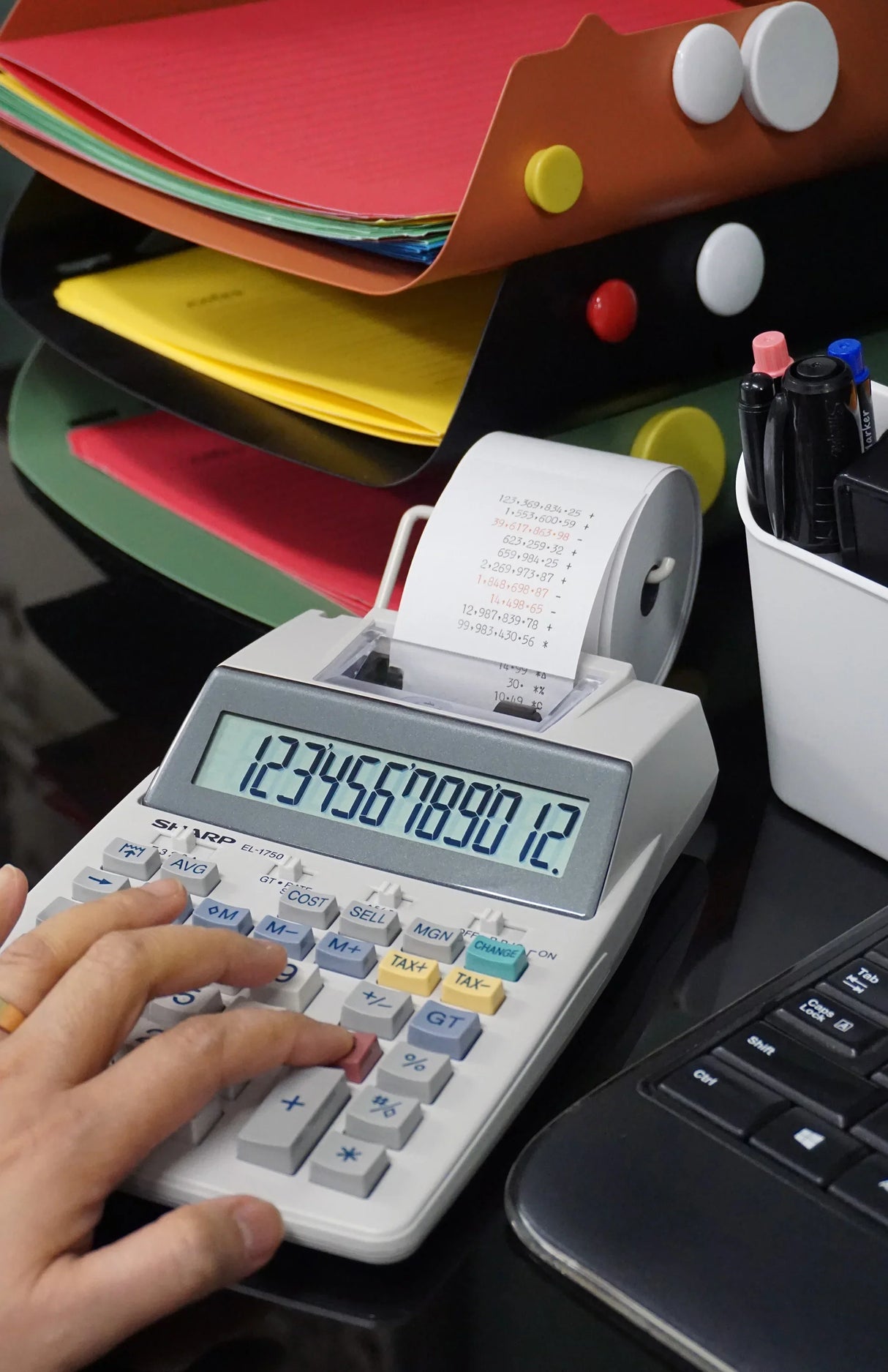 Calculator with printing paper on office desk near folders, pens, and a keyboard
