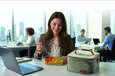 Woman eating lunch at a desk with a cityscape view, next to a Heetz heated lunch bag.
