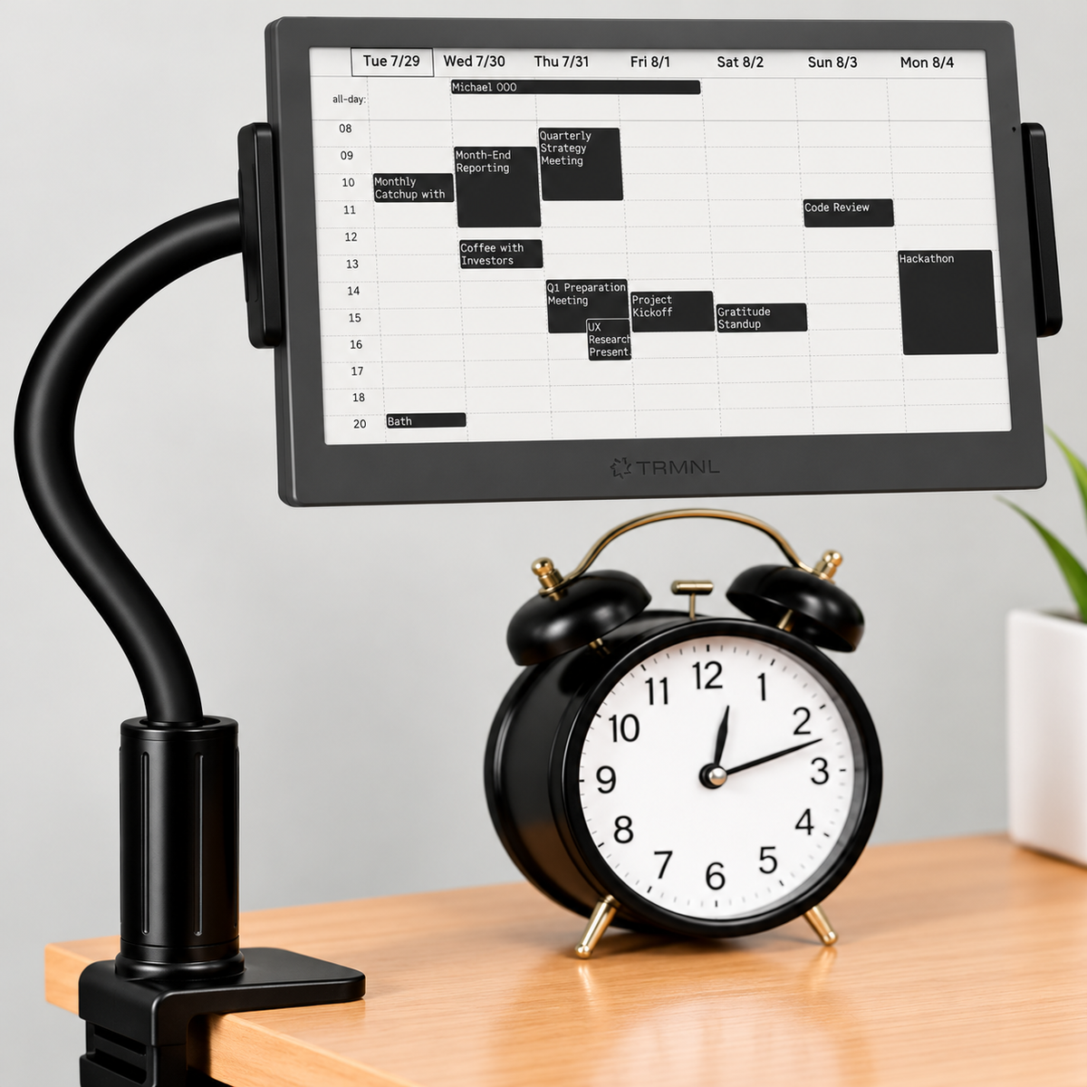 TRMNL 7.5 inch ePaper display on a gooseneck clamp next to a black alarm clock on a desk