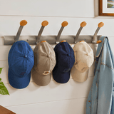 A wall-mounted metal coat rack with six wooden-handled hooks hanging four baseball caps in blue, beige, navy, and cream, with a light blue denim jacket draped over the right hook against a white paneled wall.