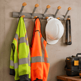 A gray metal wall-mounted coat rack with six wooden pegs hanging workwear and tools against a concrete wall, including a high-visibility yellow-green jacket, an orange safety vest, a white hard hat, and a black tool case.