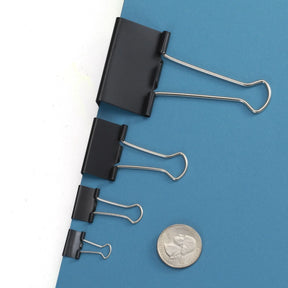 Four black Officemate binder clips of varying sizes on a blue background with a US quarter.