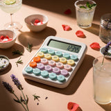Pastel desktop calculator with large buttons on a tan table with drinks and flower petals